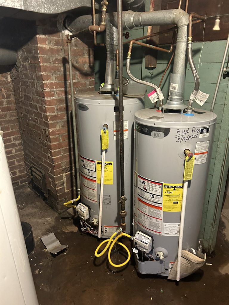 Two residential gas water heaters installed side-by-side in a basement, connected to yellow flexible gas lines and galvanized venting. Labeled "3rd Floor 3/10/2021", the units show EnergyGuide labels and digital gas meters. Visible brick wall and utility piping surround the setup.